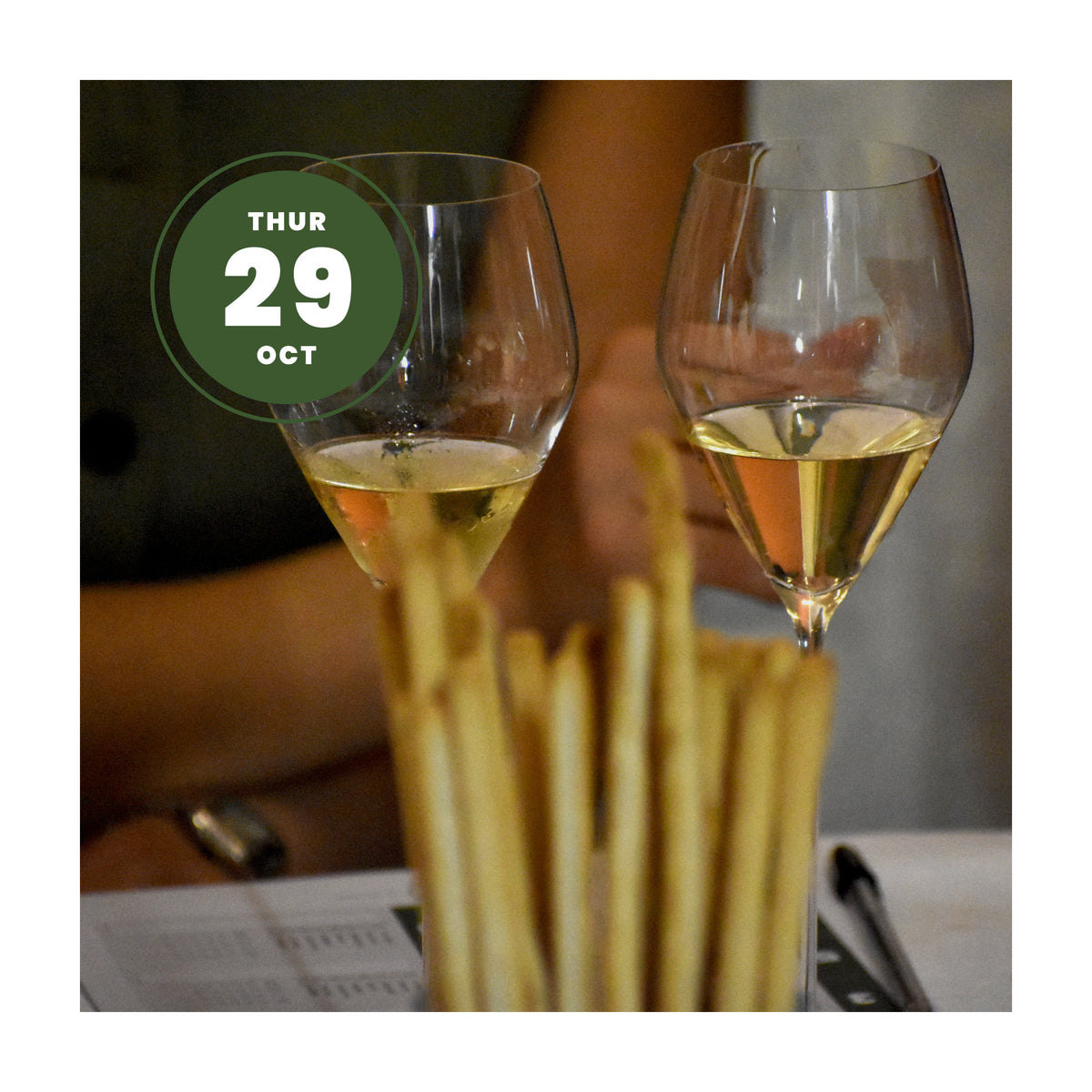 Two clear wine glasses filled with white wine, with breadsticks in the foreground and a green circular date overlay reading 'THUR 29 OCT'.