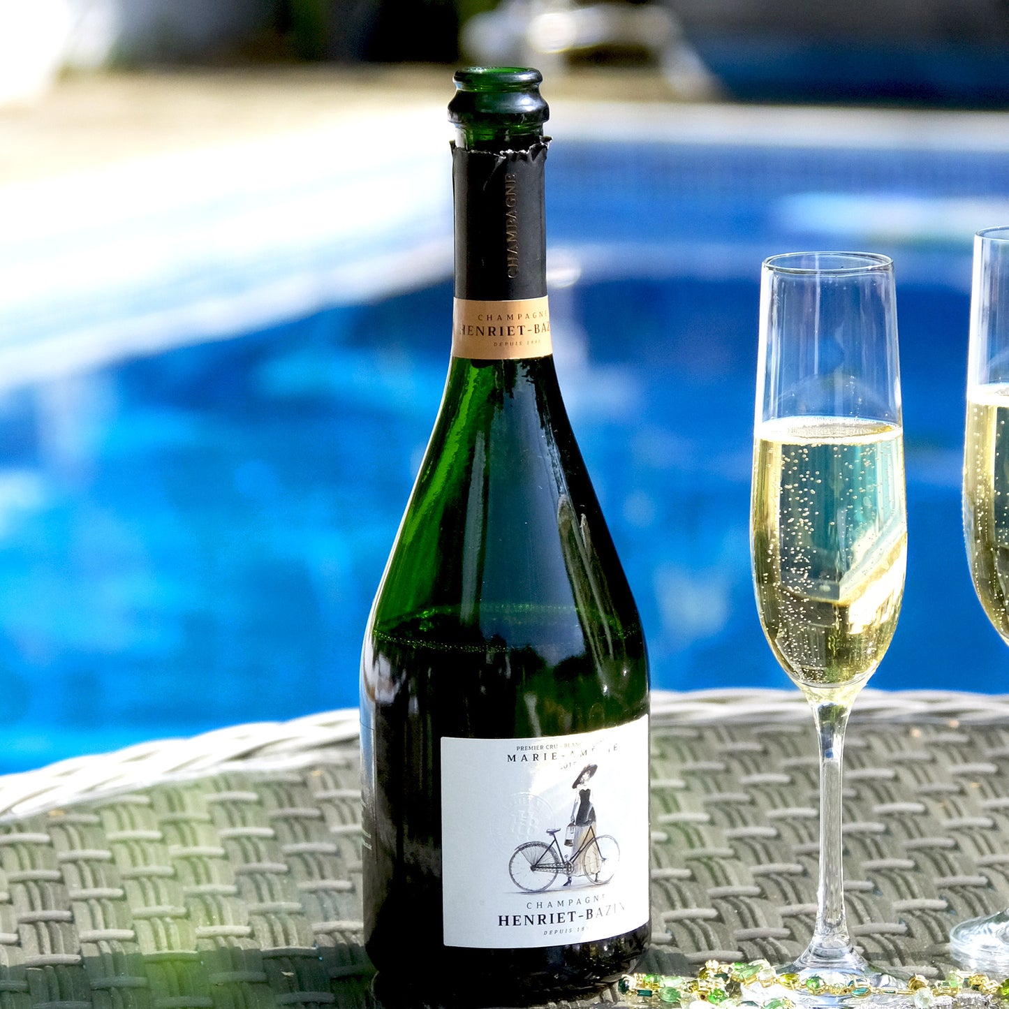 Bottle of champagne with a woman with a bike on the label and two filled glasses by a poolside