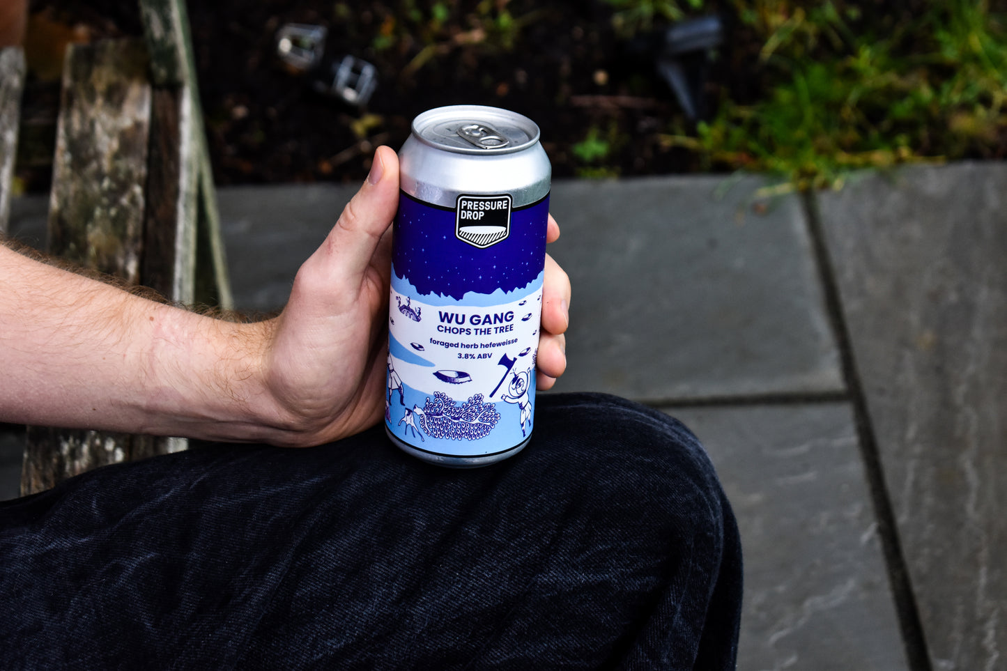 Person holding a Wu Gang beer can outdoors on a bench with flagstones