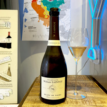 Bottle of William Saintot 100% Pinot Noir Champagne with a glass on a wooden surface, with a map and neon lights in the background.