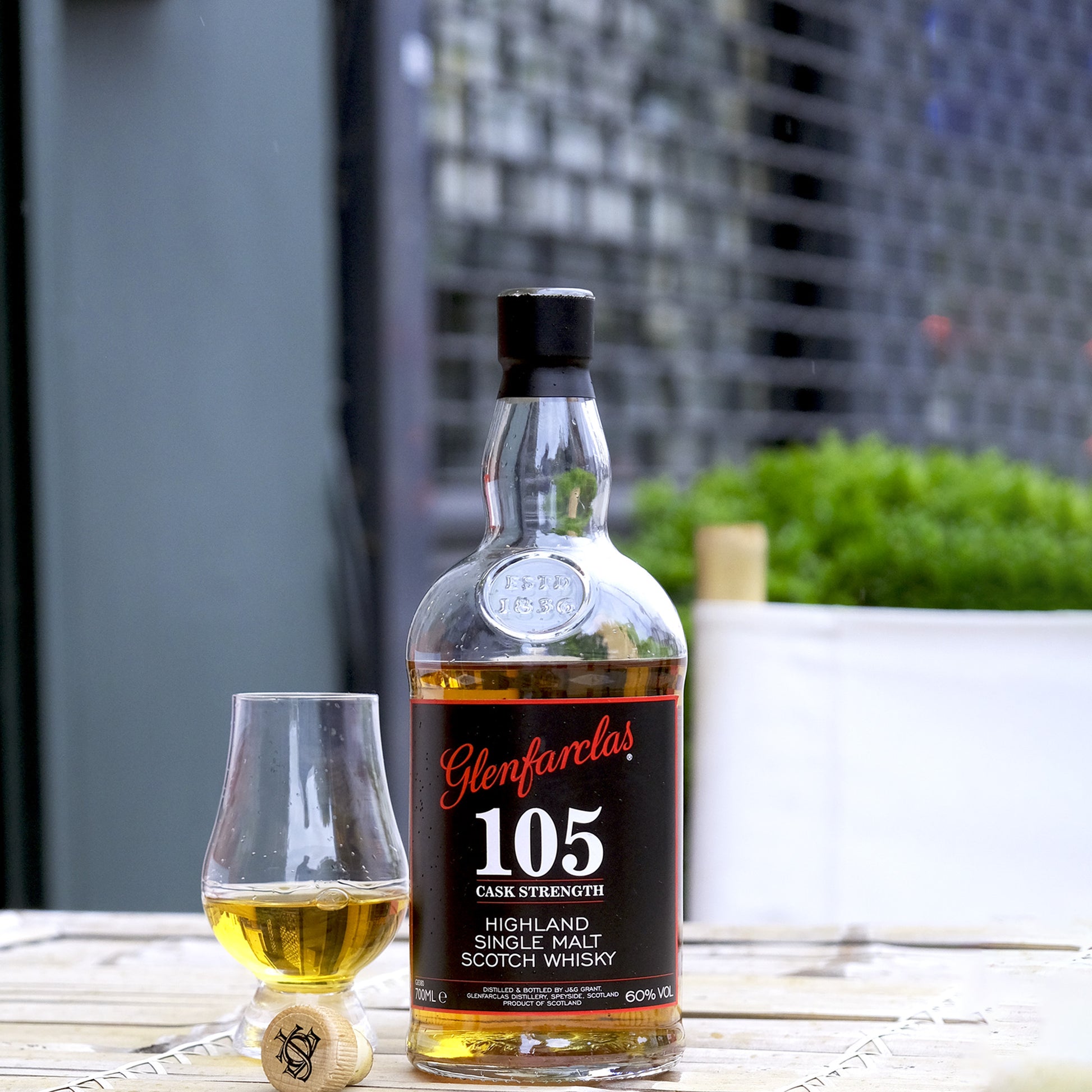 Glenfarclas 105 Cask Strength Whisky Bottle Lifestyle Shot on a table next to a glencairn glass with whisky inside in an outdoor setting.